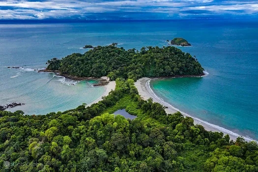 Aerial view of a lush green island connected by a sandbar between two tropical beaches, with clear blue ocean surrounding it.